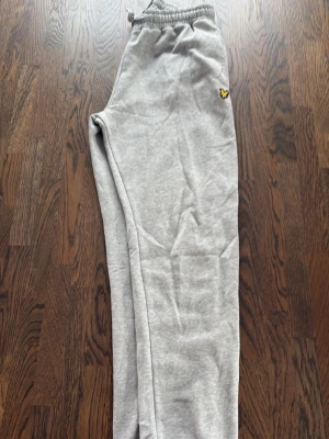 Grå mjukisbyxor från Lyle & Scott - Säljer ett par grå mjukisbyxor från Lyle & Scott med elastisk midja och en bakficka. Byxorna har det klassiska gula logomärket på vänster sida och är gjorda i mjukt bomullsmaterial. Perfekta för chill dagar eller när du vill vara bekväm.