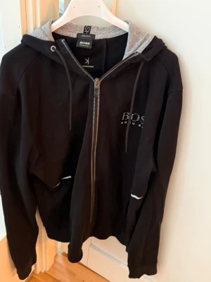 Svart zip-hoodie från BOSS - Snygg svart hoodie från BOSS Hugo Boss med dragkedja framtill och grå insida på huvan. Regular fit-modell med vita detaljer vid fickorna och logga på bröstet. Perfekt för en chill och stilren look.