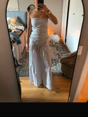 Randig axelbandslös långklänning - Säljer en snygg axelbandslös långklänning med smala vertikala ränder i vitt och grått. Klänningen har en figurnära överdel och ett långt, flödande kjolparti. Perfekt för sommarens alla tillfällen!