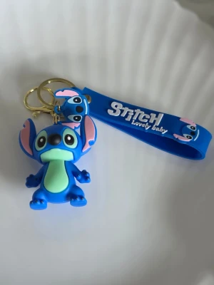 Blå Stitch nyckelring med band - Nyckelring med en blå Stitch-figur i mjukt gummi och ett matchande blått band med texten 'Stitch Lovely baby'. Perfekt att hänga på väskan eller nycklarna för en lekfull touch.