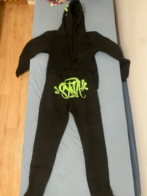Svarta syna world tracksuit - Svart tracksuit från syna world med grön graffiti-logga framtill. Byxorna har slim passform, dragkedja vid benslut och är gjorda i mjukt bomullsmaterial. Perfekta för chill eller streetwear. Kommer i cool förpackning med grönt mönster.