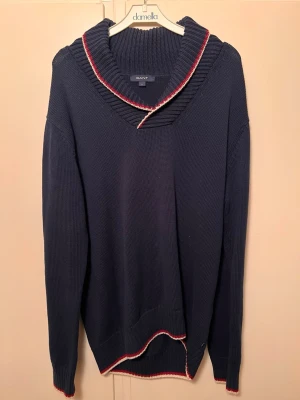 Mörkblå stickad tröja från Gant - Mörkblå stickad tröja från Gant med bred ribbad krage och snygga röda och vita detaljer längs kragen och nederkanten. Tröjan har lång ärm och avslappnad passform, perfekt för en chill och stilren look. Nypris: 3500kr