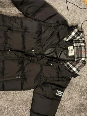 Svart dunjacka från Burberry - Snygg svart dunjacka från Burberry med klassiskt rutigt foder i huvan och tydlig logga på ärmen. Jackan har dragkedja och knappar framtill samt praktiska fickor. Perfekt för kalla dagar och riktigt stilren look. Går såklart och få jackan till en väst också