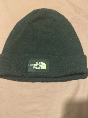 Svart mössa från The North Face - Svart ribbstickad mössa från The North Face med klassisk logotyp framtill. Enkel och stilren design som passar perfekt till kalla dagar. Tillverkad i mjukt material för extra komfort.