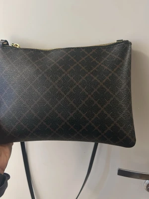 Kuvertväska  - Snygg svart axelväska från Ralph Lauren med diskret rutmönster i mörkbrunt. Väskan har guldfärgad dragkedja och justerbar axelrem. Tillverkad i tåligt konstläder med beige innerfoder. Perfekt för dig som gillar stilrena accessoarer.