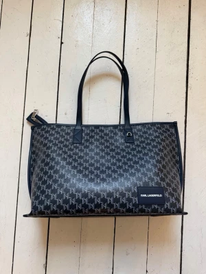 Marinblå handväska från Karl Lagerfeld - Snygg svart handväska från Karl Lagerfeld med monogrammönster i vitt och marinblå handtag. Väskan har en rektangulär form, dragkedja upptill och en logotypplatta framtill som går att ta av. Tillverkad i konstläder med en stilren och modern look. Ny pris runt 3000kr!