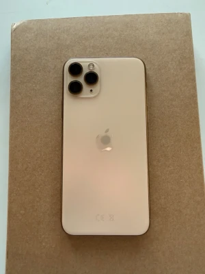 iPhone 11 Pro guld med trippelkamera - iPhone 11 Pro i guldig färg med snygg glasbaksida och tre kameror på baksidan. Telefonen har en stor skärm med tunna kanter och rundade hörn. Apple-loggan syns tydligt på baksidan och mobilen känns riktigt premium med sin stilrena design.