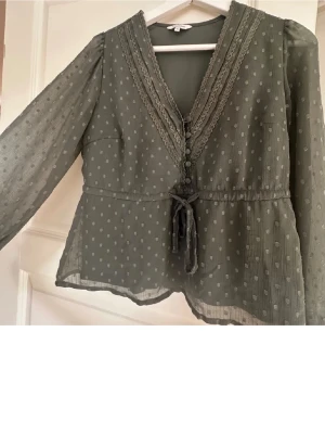 Olivgrön blus med spets från Bubbleroom - OBS!! HAR EN VIT FÅRG FLÄCK LÄNGST NED PÅ ÄRMEN PRECIS DÄR HANDEN ÄR SKICKAR BILD PRIVAT PÅ DEN!! Snygg olivgrön blus från Bubbleroom med prickigt mönster, v-ringning med spetsdetaljer och knytband i midjan. Blusen har genomskinliga långärmade ärmar och dekorativa knappar framtill. Perfekt för dig som gillar romantiska detaljer och en avslappnad men chic look.