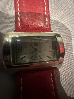 Röd klocka från Faded Glory - Snygg klocka från Faded Glory med rektangulärt silverfärgat ur och röd rem med vita sömmar. Klockan har en klassisk analog urtavla och ett modernt, lite kantigt utseende. Remmen är i skinnimitation och urtavlan har tydliga siffror vid 3, 6, 9 och 12. Funkar ej!