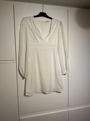 Vit kortklänning med v-ringning - Snygg vit kortklänning med v-ringad hals och långa ärmar. Klänningen har en feminin siluett och stängs med dragkedja bak. Perfekt för dig som gillar en stilren och fräsch look.