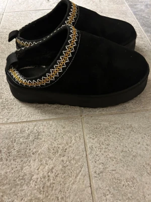 Svarta uggs  med broderad kant - Säljer ett par svarta tofflor i mjukt material med broderad kant i gult och vitt. Tofflorna har en tjock sula och ögla bak för enkel påtagning. Perfekta för att chilla hemma och hålla fötterna varma. Sulan innuti är lite av, man märker ej det när man använder dem då det bara är tyg sula men går att lägga in en ny. 