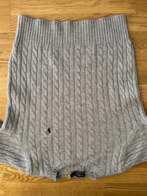 Grå kabelstickad tröja Polo Ralph Lauren - Kabelstickad grå tröja från Polo Ralph Lauren med rund halsringning och ribbade muddar. Tröjan har ett broderat svart logomärke på bröstet och är tillverkad i mjukt material. Perfekt för lager-på-lager-stil under kyliga dagar.