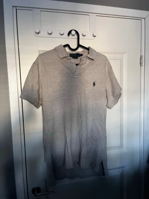 Grå pikétröja från Ralph Lauren - Klassisk beige pikétröja från Ralph Lauren med broderad logga på bröstet. Tröjan har krage, knappar framtill och korta ärmar. Perfekt för en stilren och avslappnad look.