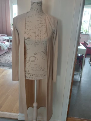 Beige lång ribbad kofta - Säljer en beige lång kofta med ribbad struktur och öppen front. Koftan har långa ärmar och är tillverkad i ett mjukt, stretchigt material som faller snyggt längs kroppen. Perfekt att slänga över en topp för en avslappnad look.