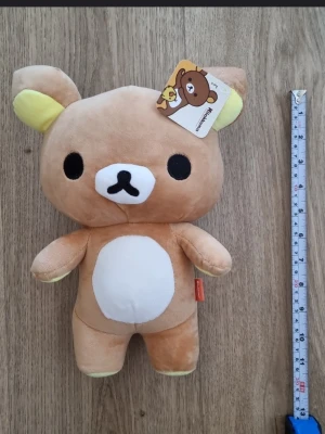 Rilakkuma mjukisdjur med dragkedja - Supergulligt Rilakkuma mjukisdjur från San-X, ca 32 cm långt. Nallen är ljusbrun med vita och gula detaljer, rund mage och svart broderat ansikte. Baksidan har en dragkedja och liten svans. Tillverkad i mjuk plysch och perfekt som samlarobjekt eller dekoration.