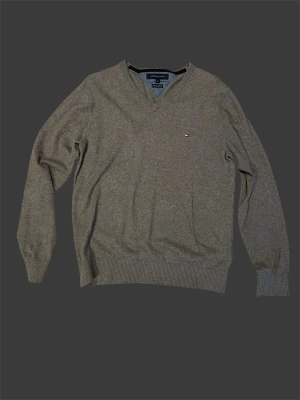 Grå stickad tröja Tommy Hilfiger - Klassisk grå-brun stickad tröja från Tommy Hilfiger med v-ringning och diskret logga på bröstet. Tröjan har långa ärmar och ribbade muddar vid ärmslut och nederkant. Perfekt för lager på lager och enkel att matcha med olika outfits.