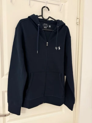 Marinblå hoodie från Polo Ralph Lauren - Snygg marinblå hoodie från Polo Ralph Lauren med dragkedja framtill, två fickor och klassisk vit logga på bröstet. Huva med dragsko och ribbade muddar. Tillverkad i mjuk bomullsblandning som känns riktigt skön mot huden.