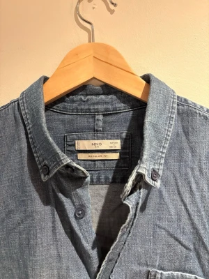 Blå jeansskjorta från MNG - Säljer en klassisk blå jeansskjorta från MNG med regular fit. Skjortan har lång ärm, bröstficka och knappar framtill. Perfekt till ett avslappnat men ändå snyggt uttryck. Tillverkad i mjukt bomullsmaterial.