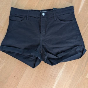 Svarta shorts från H&M, strl 38 - Svarta shorts från H&M i klassisk femficksmodell med uppvikta benslut. De har knapp och dragkedja framtill samt bälteshällor. Materialet är mjuk bomull med lite stretch för skön passform. Perfekta för varma dagar och enkel att matcha med din favorit-topp.