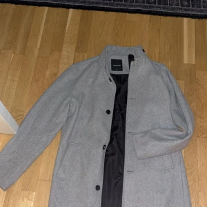 Grå rock från jack n jones - Snygg grå rock från Jack n Jones med klassisk rak passform och svarta knappar. Jackan har ståkrage, två sidofickor och är helfodrad med svart innerfoder. Perfekt för dig som vill ha en stilren look under höst och vinter.