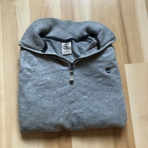 Grå half zip tröja från Timberland - Snygg grå långärmad tröja från Timberland med half zip och ribbad krage. Tröjan har ett diskret svart Timberland-logo broderat på bröstet och är tillverkad i mjuk bomull. Perfekt för dig som gillar stilren och enkel design.