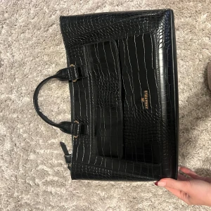 Svart handväska i croco-look från Anna Field - Snygg svart handväska från Anna Field med croco-mönstrad yta och guldfärgade detaljer. Väskan har två handtag, dragkedja upptill och rymligt innerfack. Perfekt för dig som vill ha en stilren och trendig accessoar med lyxig känsla. Finns en axelrem till