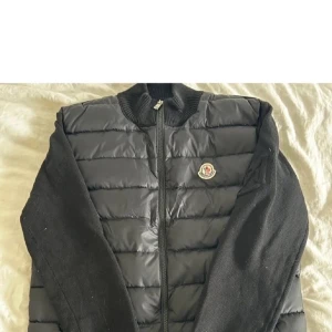 Svart Moncler pufferjacka med stickade ärmar - Säljer en svart pufferjacka från Moncler med stickade ärmar och quiltad front. Jackan har Moncler-logga på bröstet och dragkedja framtill. Materialet är en mix av dunfyllning och stickad polyester, perfekt för dig som gillar stilren och sportig look.