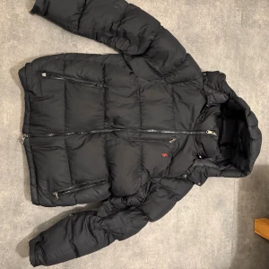 Svart pufferjacka från Polo Ralph Lauren - Svart pufferjacka från Polo Ralph Lauren med huva, flera dragkedjeförsedda fickor och diskret röd logga på bröstet. Jackan har quiltad design och är tillverkad i syntetmaterial, perfekt för kalla dagar. Klassisk look med normal passform och långa ärmar.