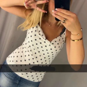 Vit prickig linnetopp med draperad hals - Säljer en vit linnetopp med svarta prickar och draperad halsringning. Toppen är ärmlös med smala axelband och har en lätt, mjuk känsla. Perfekt att styla med jeans eller kjol för en fräsch look. Passar dig som gillar klassiska mönster med en twist.