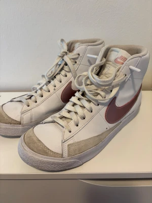 Nike Blazer Mid vit/rosa EUR 40 - Nike Blazer Mid sneakers i vitt skinn med ljusrosa metallic swoosh och broderad Nike-logga på hälen. Klassisk high-top modell. Grå mockadetaljer vid tå och sidor. Har knappt använt. Köptes för 700 kr. Priset kan diskuteras.