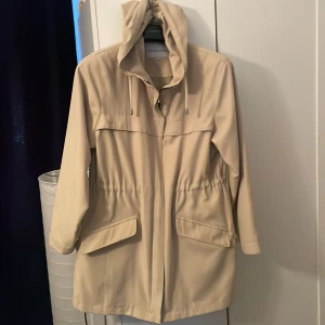 Beige parkajacka från Exlindexlin - Snygg beige parkajacka från Exlindexlin med huva och snörning. Jackan har långa ärmar, två stora fickor med lock och dragkedja framtill. Perfekt för dig som gillar stilren och enkel design med praktiska detaljer.