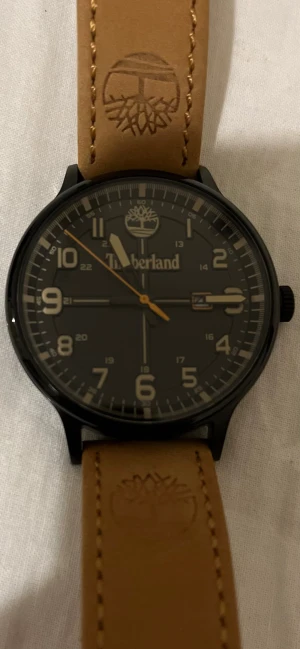 Svart Timberland klocka med läderband - Snygg Timberland klocka med svart urtavla och tydliga siffror i beige. Boetten är svart och armbandet är gjort av ljust brunt läder med Timberland-logga ingraverad. Klockan har datumvisning och detaljer i silver och orange. Perfekt accessoar för dig som gillar stilren design.