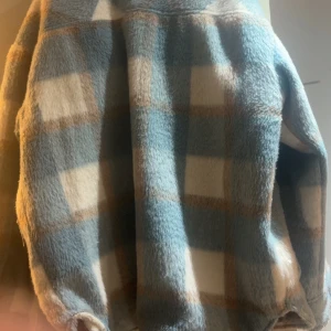 Blå och beige rutig teddyjacka - Säljer en sjukt mysig teddyjacka med rutigt mönster i blått, beige och vitt. Jackan har stora fickor framtill och knäpps med knappar. Perfekt för dig som gillar chill och oversized vibe. Materialet är fluffigt och mjukt, och jackan har lång ärm.