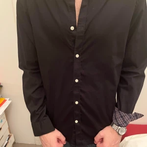 Svart skjorta med rutiga detaljer - Svart skjorta från Brooks Brothers med vita knappar och rutigt foder i krage och manschetter. Klassisk button-down krage och långärmad modell. Skjortan är i bomull och har en normal passform. Perfekt för dig som gillar stilrena plagg med en twist.