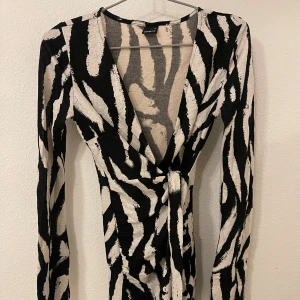 Zebramönstrad omlottklänning Gina Tricot - Supersnygg kort omlottklänning från Gina Tricot i svartvitt zebramönster. Klänningen har lång ärm, markerad midja med knyt och djup v-ringning. Perfekt för dig som vill sticka ut med en trendig look.