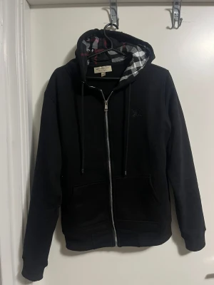 Svart Burberry hoodie med dragkedja - Svart hoodie från Burberry med klassiskt rutigt foder i huvan och broderad logga på bröstet. Tröjan har dragkedja framtill, snörning i huvan och två fickor. Perfekt för dig som vill ha en stilren och lyxig streetwear-look.