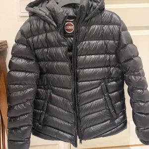 Svart Colmar dunjacka med huva - Svart dunjacka från Colmar med huva. Jackan har dragkedja framtill, två sidofickor och en snygg, glansig finish. Perfekt för kalla dagar. Mycket liten reva på framsida som knappt syns. Annars helt i nyskick!!