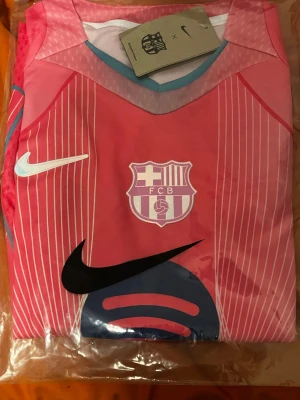 Barcelona Lamine Yamal 10 Nike fotbollströja - Säljer en Barcelona fotbollströja med Lamine Yamal och nummer 10 på ryggen. Tröjan är rosa med vita och blå detaljer, har tunna vita ränder och är kortärmad. Nike Dri-FIT material för skön känsla och bra ventilation. FCB klubbmärke och Nike-logga på bröstet. Den är helt ny