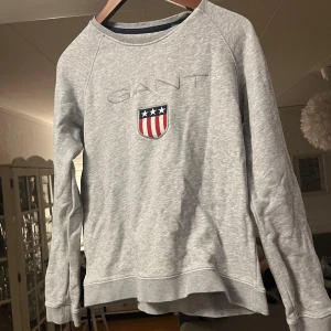 Grå sweatshirt från GANT med logga - Snygg grå sweatshirt från GANT med broderad logga och sköld på bröstet. Tröjan har rund halsringning, långa ärmar och är tillverkad i mjuk bomullsmix. Perfekt för en chill och stilren look.