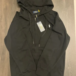 Svart hoodie från Polo Ralph Lauren - Svart hoodie från Polo Ralph Lauren med dragkedja, huva och snörning. Klassisk vit broderad logga på bröstet och ribbade muddar. Perfekt för en chill och stilren look. Tillverkad i mjuk bomull för maximal komfort.