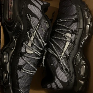 Nike Air Max Plus Utility svart/grå - Nike Air Max Plus Utility sneakers i svart och grått med reflekterande detaljer och grova snören. Ovandel i syntet och mesh med mönstrad struktur, robust platt sula med synliga Air-enheter och Vibram-yttersula för extra grepp. Swoosh-logga i silver på sidan.
