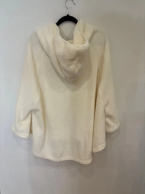 Krämvit fleece hoodie med tofsar - Supermjuk och fluffig hoodie i krämvit fleece från Crossbow. Tröjan har huva med långa snören och söta tofsar, samt vida trekvartsärmar och avslappnad passform. Perfekt för att mysa hemma eller chilla med vänner.