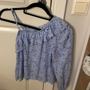 Blommig offshoulder topp från Lindex - Söt ljusblå offshoulder topp från Lindex med blommigt mönster i vitt och blått. Toppen har en volang längs halsringningen och en axelbandslös sida samt en lång ärm. Perfekt för vår och sommar när du vill ha en luftig och trendig look.