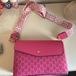 Rosa axelväska med mönstrad rem - HELT NY!!🩷( aldrig använd )Snygg rosa axelväska med bred , mönstrad axelrem i vitt och rosa. Väskan har ett M-mönster över hela tyget, guldiga detaljer och praktiska fack inuti. Perfekt för dig som vill sticka ut med färg och stil. 🩷