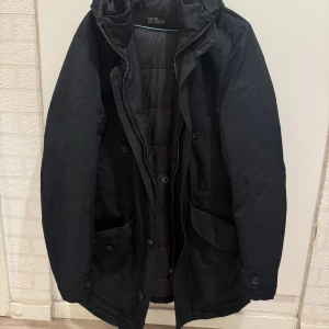 Svart parkajacka från Oscar Jacobson - Svart parkajacka från Oscar Jacobson med quiltad insida, huva och flera fickor med knappstängning. Jackan har dragkedja och knappar framtill, samt diskret logoband på insidan. Perfekt för kalla dagar med sin långa, rymliga passform. Storlek 46.