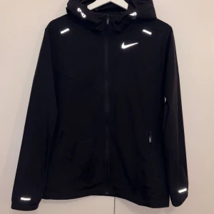 Nike Miller Windbreaker - Svart vindjacka från Nike med huva och hel dragkedja framtill. Jackan har reflekterande detaljer och elastiska muddar vid ärmsluten. Perfekt för blåsiga dagar och sportiga outfits. Swoosh-logga på bröstet ger en clean och sportig look.