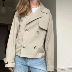 Beige kort trenchcoat - Snygg kort trenchcoat i beige med dubbelknäppning och stora knappar. Jackan har klassisk krage, bältesdetaljer vid ärmsluten och en rak, loose passform. Perfekt för dig som vill ha en stilren och trendig jacka till vardags.