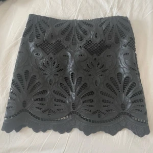 Svart kortkjol med spetsdetaljer - Snygg svart kortkjol med unikt spetsmönster och vågig kant. Kjolen har ett coolt, genombrutet designmönster som ger en edgy vibe. Perfekt för dig som gillar statement-plagg och vill sticka ut med detaljerad look. Har endast använts 1 gång och är precis som ny 💕