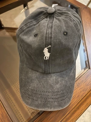 Svart keps från Polo Ralph Lauren - Snygg svart keps från Polo Ralph Lauren med klassisk broderad logga framtill och 'POLO' broderat baktill. Kepsen har böjd skärm, justerbar rem med metallspänne och är tillverkad i mjuk bomull. Perfekt för en chill och sportig look.