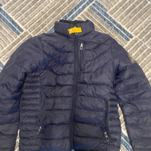 Mörkblå dunjacka från Parajumpers - Säljer en mörkblå dunjacka från Parajumpers med gul detalj vid halsen och snygga silverfärgade dragkedjor. Jackan har quiltad design, hög krage och långärmade ärmar. Märkespatch på vänster ärm. Perfekt för kalla dagar. Är sliten på några platser kan skicka bild vid önskemål 